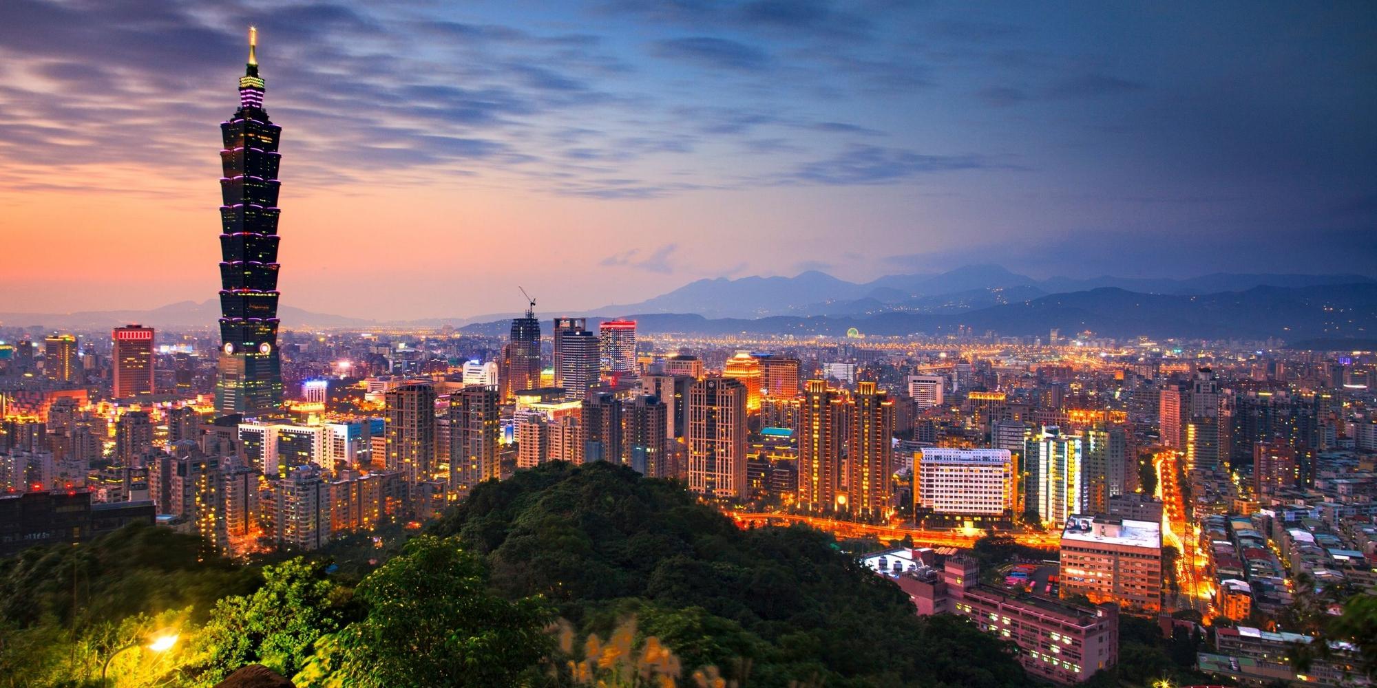 Taipei skyline lit at dusk, seen from a wooded hillside with mountains behind the city
