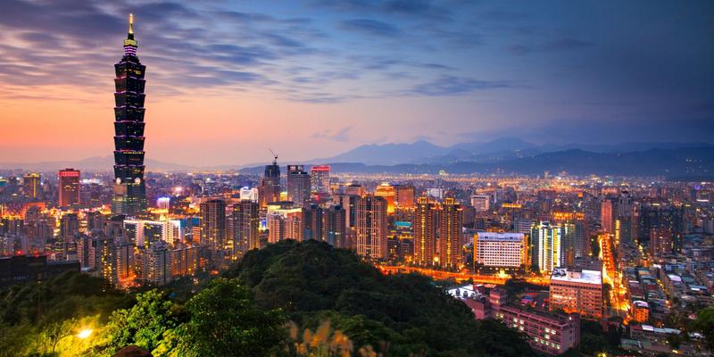 Taipei skyline lit at dusk, seen from a wooded hillside with mountains behind the city